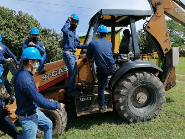 Estudiantes con casco suben a una retroexcavadora durante una práctica de maquinaria amarilla